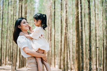 Fototapeta premium Asian mother carrying a daughter in arms in a peaceful forest setting, Happy smile mother and little child playing together outdoor, Family day memory on holiday concept