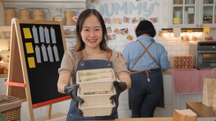 Smiling woman in an apron holding food containers in a cozy kitchen environment, promoting takeout meal services.