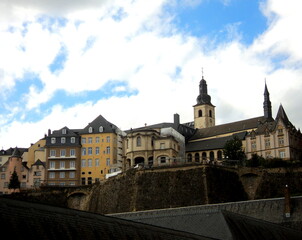 Naklejka premium A picturesque view of Luxembourg's Grund, with historic stone buildings, winding cobblestone streets, and lush greenery lining the river beneath towering cliffs.
