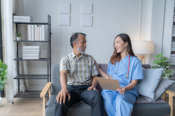 Young female nurse wearing blue scrubs is holding a clipboard and smiling at an elderly man sitting on a sofa in his living room