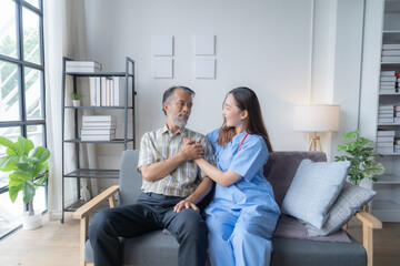 Young nurse wearing blue scrubs is comforting a senior patient while sitting on a couch in a home. The nurse is holding the patient's hand and they are both smiling at each other