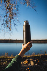 Obraz premium A thoughtful individual is gently holding a thermos while sitting beside a serene lake, fully immersed in the stunning view and the calming atmosphere of nature surrounding them