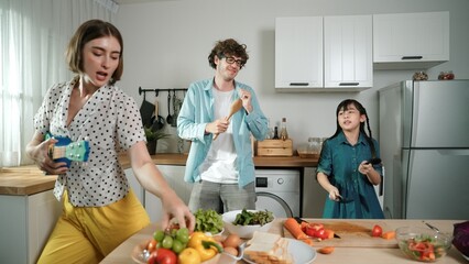 Caucasian skilled father, mother and asian daughter making breakfast while dancing together. Skilled mom playing ukulele while preparing vegetable at modern kitchen. Healthy food concept. Pedagogy.