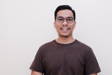 A Southeast Asian man wearing brown tshirt and glasses cream background happy face smiling.