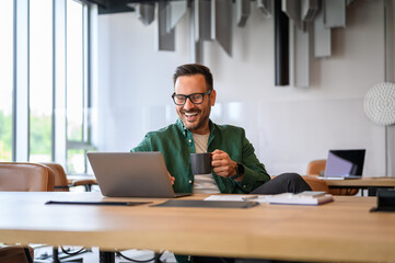 Cheerful male engineer holding coffee cup and working on project online over laptop at desk in...