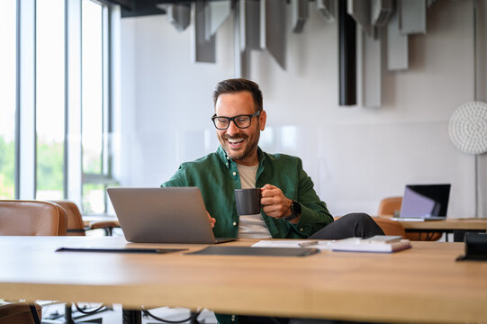 Cheerful male engineer holding coffee cup and working on project online over laptop at desk in office