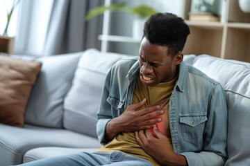 Man suffers from heart attack on gray couch. Chest pain, clutching hand, yellow shirt, blue denim jacket. Background plant, window, wall. Severe health condition, emergency, stress, fear.