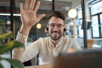 Fototapeta premium Young businessman talks on video call at office desk. Man smiles, waves at smartphone camera, break from work. Businessman holds pen, sits with laptop, plant. Indoor setting with window, blinds.