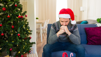 Sad man wearing a santa hat feeling depressed and lonely during christmas holidays sitting on the...