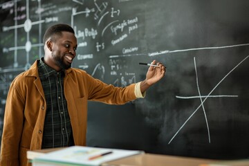 African American man in classroom stands in front of blackboard. He points to equation on whiteboard with marker. Smiling face shows sense of accomplishment. Lesson or presentation takes place.
