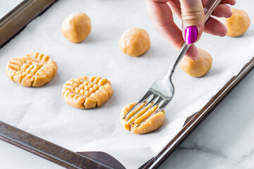 A hand pressing a fork into raw peanut butter cookies to make cross hatches.