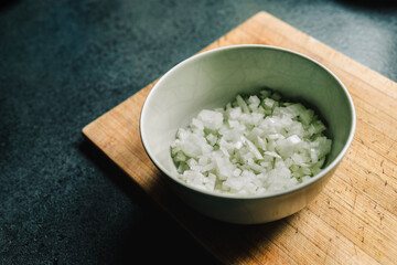 Finely diced onion in a beige bowl on a wooden used cutting board. Dark kitchen countertop