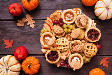 Autumn theme dessert board. Top view table scene against a dark wood background. Pumpkin, apple and pecan mini pies, cookies, pumpkin rolls and fruit. © Jenifoto