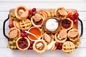 Fall pie theme dessert board. Overhead view table scene against a white wood background. Pumpkin, apple and pecan mini pies, with whipped cream, caramel and fruit.