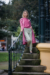 A woman in a pink coat stands on stone steps in a quiet park, the autumn air cool and the overcast sky creating a peaceful, contemplative mood.