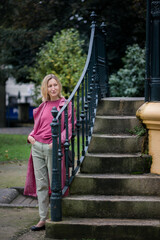 A woman in a pink coat stands beside the stone steps in a park, the cool autumn air a peaceful, reflective atmosphere.