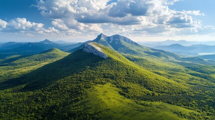 Fototapeta premium Aerial view of a mountain range with lush green forests and a clear blue sky with white clouds.