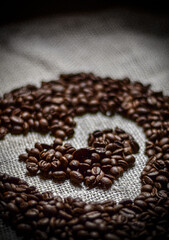 The image shows a close-up of dark, roasted coffee beans. One bean is in focus, highlighting its natural seam. The rest are blurred, creating a rich, textured background.

