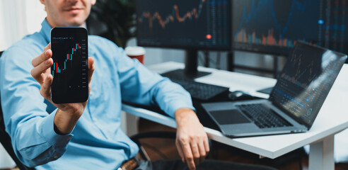 Successful young business trader showing dynamic stock market graph on smartphone, presenting infomation database of currency exchange rate in real time against on pc blurred background. Gusher.