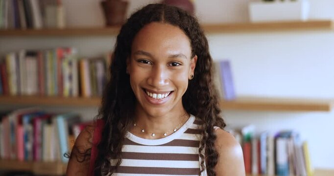 Serious beautiful long curly haired teenage African student girl posing in public university library standing with bookshelves in background, getting happy, looking at camera. Head shot portrait