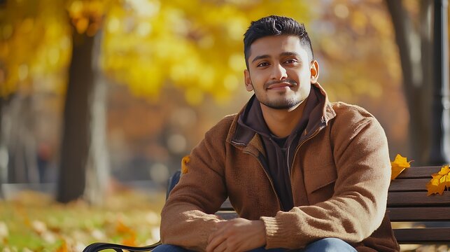 A portrait of a South Asian man sitting on a park bench with autumn leaves scattered around.