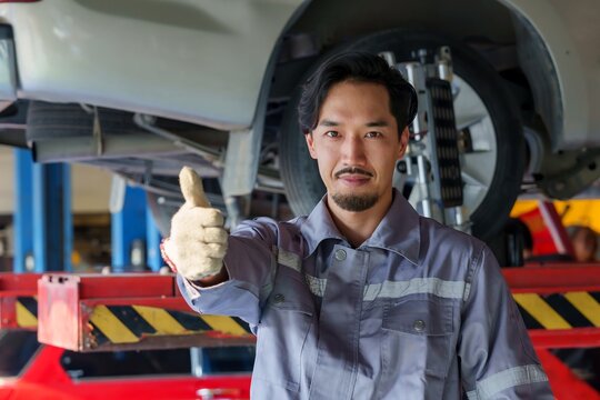 Male mechanic in grey uniform showing thumbs up while standing in front of raised vehicle in auto repair shop. confident and satisfied, expressing approval of completed job in professional environment
