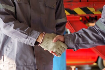 Two male mechanics shaking hands in a repair shop. Wearing grey uniforms with reflective strips, demonstrating teamwork and collaboration in a professional automotive environment.