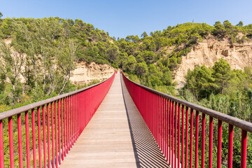 Obraz premium Puente colgante con barandillas rojas sobre el río Gállego, rodeado de vegetación y montañas, bajo un cielo despejado.
