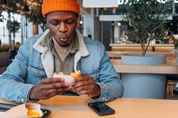 African American man eating burger with fries at table in catering restaurant on mall food court, relaxed atmosphere, urban lifestyle, smartphone on table