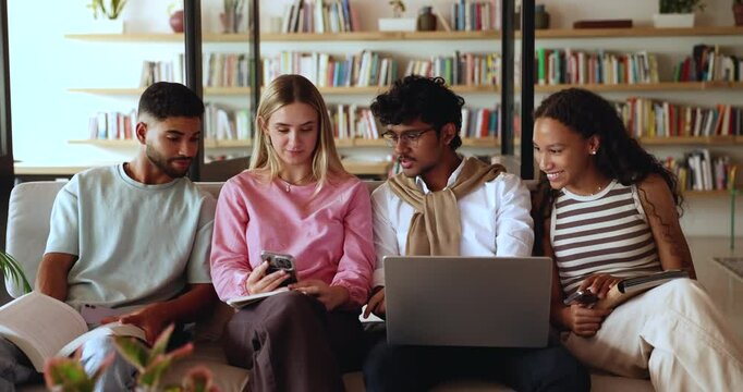 Diverse group of fresh students discussing application on smartphone, looking at gadget on classmate girl, sitting on couch together, talking, using digital devices in campus library space