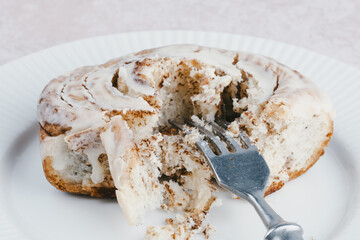 Close up a cinnamon roll sits on a white plate, with a fork resting beside it, showcasing its spiraled pastry and creamy frosting. The cozy kitchen adds warmth to the scene.