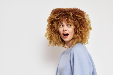 Woman with curly hair, surprised expression, wearing a light blue sweater against a plain white background, showcasing emotions and healthy hair