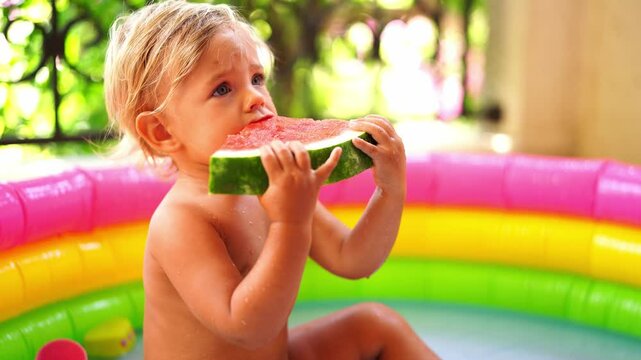 Little girl nibbles on a piece of watermelon while sitting in an inflatable pool on the balcony. High quality 4k footage