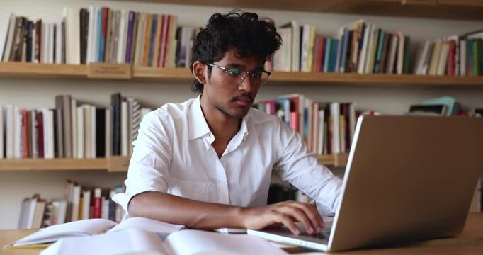 Serious successful Indian student guy in glasses typing on laptop in library, reading open book, textbook, notes, summary, preparing report for class, working on research study project