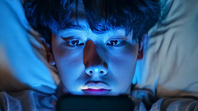 An Asian teenage boy lies in bed at night with the glow of his phone illuminating his face. He wears a neutral-colored t-shirt, and his expression is serene yet contemplative. The scene suggests