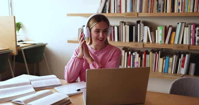 Positive young Caucasian high school student girl in headphones talking on video conference chat at laptop, waving greeting hand hello, using Internet communication for education