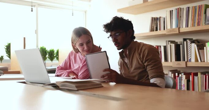 Young Indian tutor man training Caucasian student girl in library, explaining learning notes. Two diverse college mates working on research study, doing home assignment at laptop together, talking