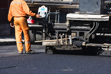 A construction worker in bright orange attire operates an asphalt paving machine on a road in...