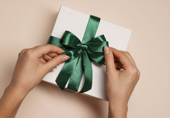 Woman decorating gift box with bow on beige background, top view