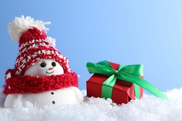 Cute decorative snowman and gift box on artificial snow against light blue background, closeup