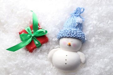 Cute decorative snowman and gift box on artificial snow, flat lay