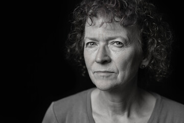 Black and white portrait of a middle aged woman with curly hair, looking at camera with a serious expression. Off centre, room for copy. 