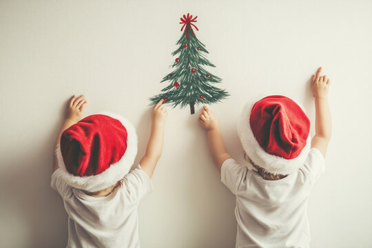 Little twins in Santa hats joyfully glue decorations to a festive Christmas tree drawing on the wall in a cozy home setting - Powered by Adobe