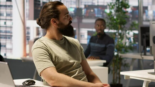 Worker sitting at office desk using computer during job shift, being interrupted by manager to receive info. Company employee in workspace doing tasks, listening to feedback from his boss, camera A