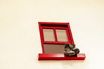 A cat is sitting on a pair of shoes that are placed on a ledge next to a red window. The shoes are old and worn, and the cat seems to be enjoying its time lounging on them. The window is old style