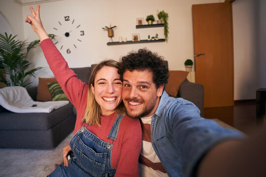 Joyful young adult Caucasian couple taking selfie looking smiling at camera at home. Cheerful people in love posing hugging mobile photo in living room. Millennial happy man and woman and social media
