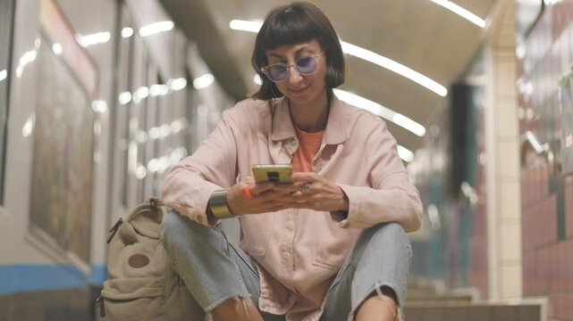 Young woman waiting for train at station and using her phone. watch schedule. Traveling by public transport.  Young girl in stylish clothes with backpack in subway. 