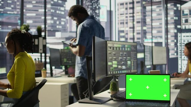Green screen laptop on desk in workspace with admins in background doing software quality assurance. Chroma key notebook in office with woman at work inspecting coding on device, camera B
