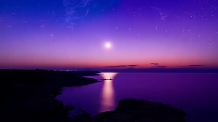 The full moon illuminates a tranquil coastline where the calm ocean stretches into the horizon. The sky is painted in soft shades of purple and pink creating a peaceful almost surreal seascape.