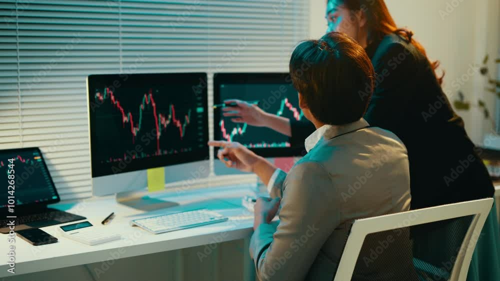 Poster Businesswoman pointing at computer monitors displays stock market data while explaining insights to her male colleague in a collaborative office setting